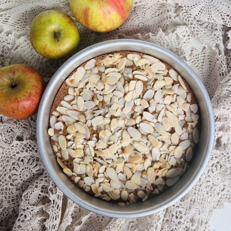 A round cake topped with sliced almonds sits in a metal baking pan on a lace cloth. Three apples are placed beside the pan, adding a rustic touch to the scene.