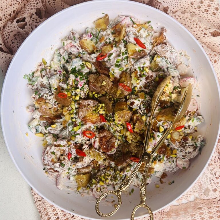 A bowl of creamy potato salad topped with crispy fried onions, sliced red chilies, and chopped herbs. Elegant gold-toned serving tongs lie atop the salad, and a lacy pink tablecloth partially covers the table beneath the bowl.