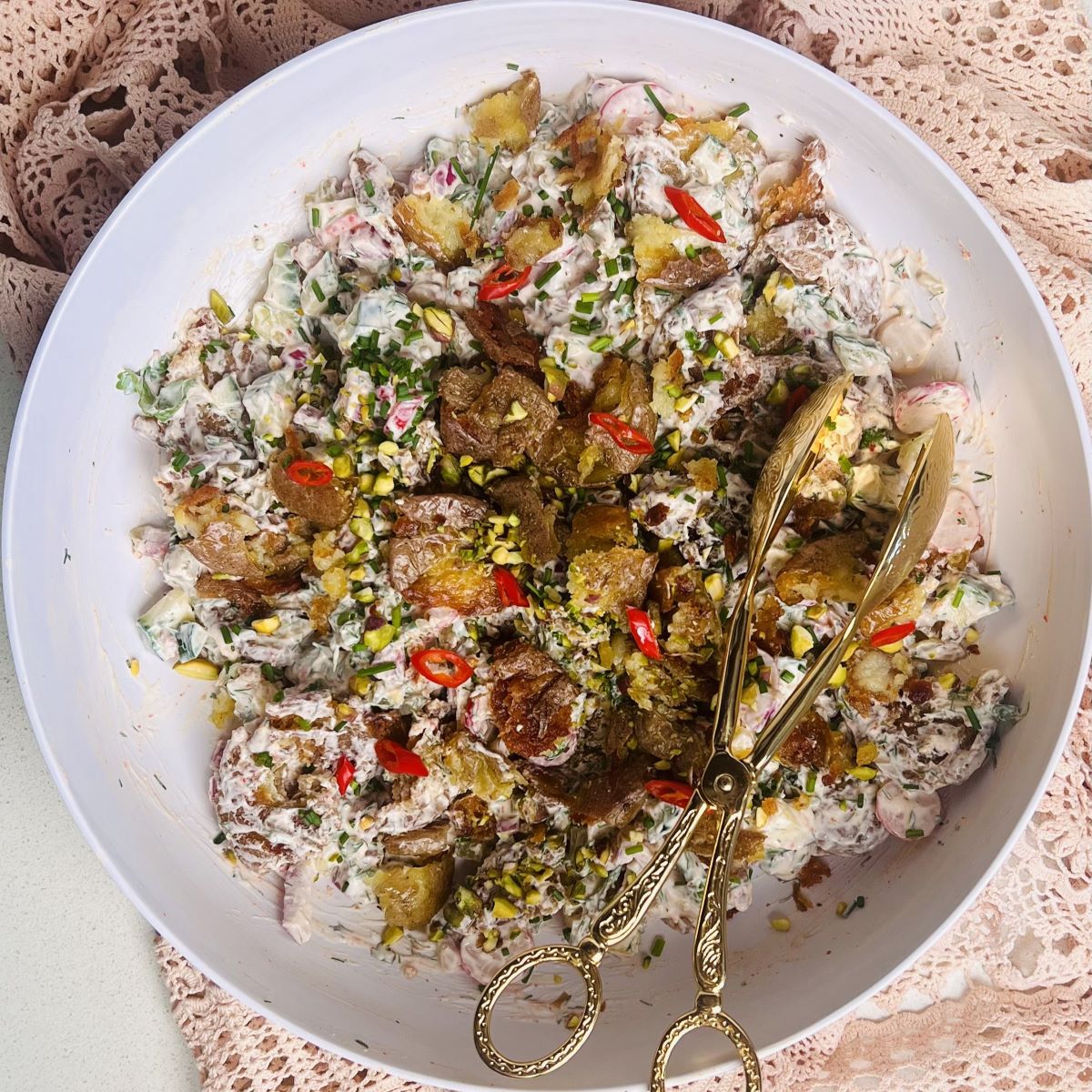 A bowl of creamy potato salad topped with crispy fried onions, sliced red chilies, and chopped herbs. Elegant gold-toned serving tongs lie atop the salad, and a lacy pink tablecloth partially covers the table beneath the bowl.