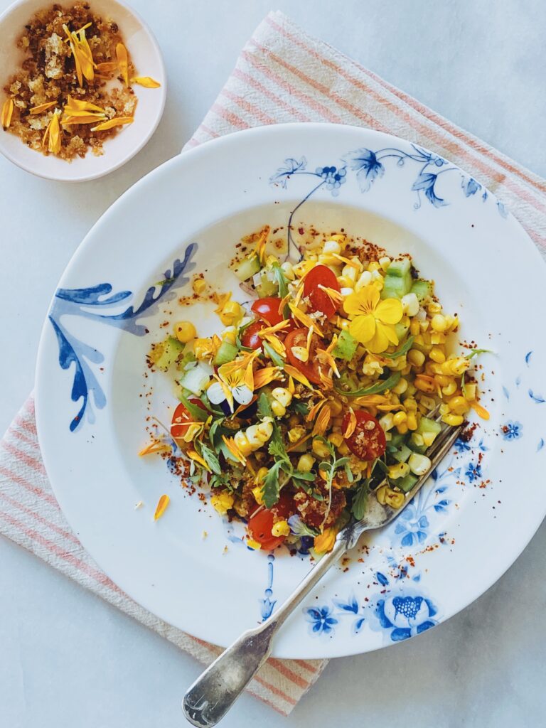 A colorful salad with cherry tomatoes, corn, cucumber, greens, and edible flowers is arranged on a white plate with blue designs. A fork and striped napkin are nearby, paired perfectly with crisp sweetcorn pakoras and a small bowl of seasoning.